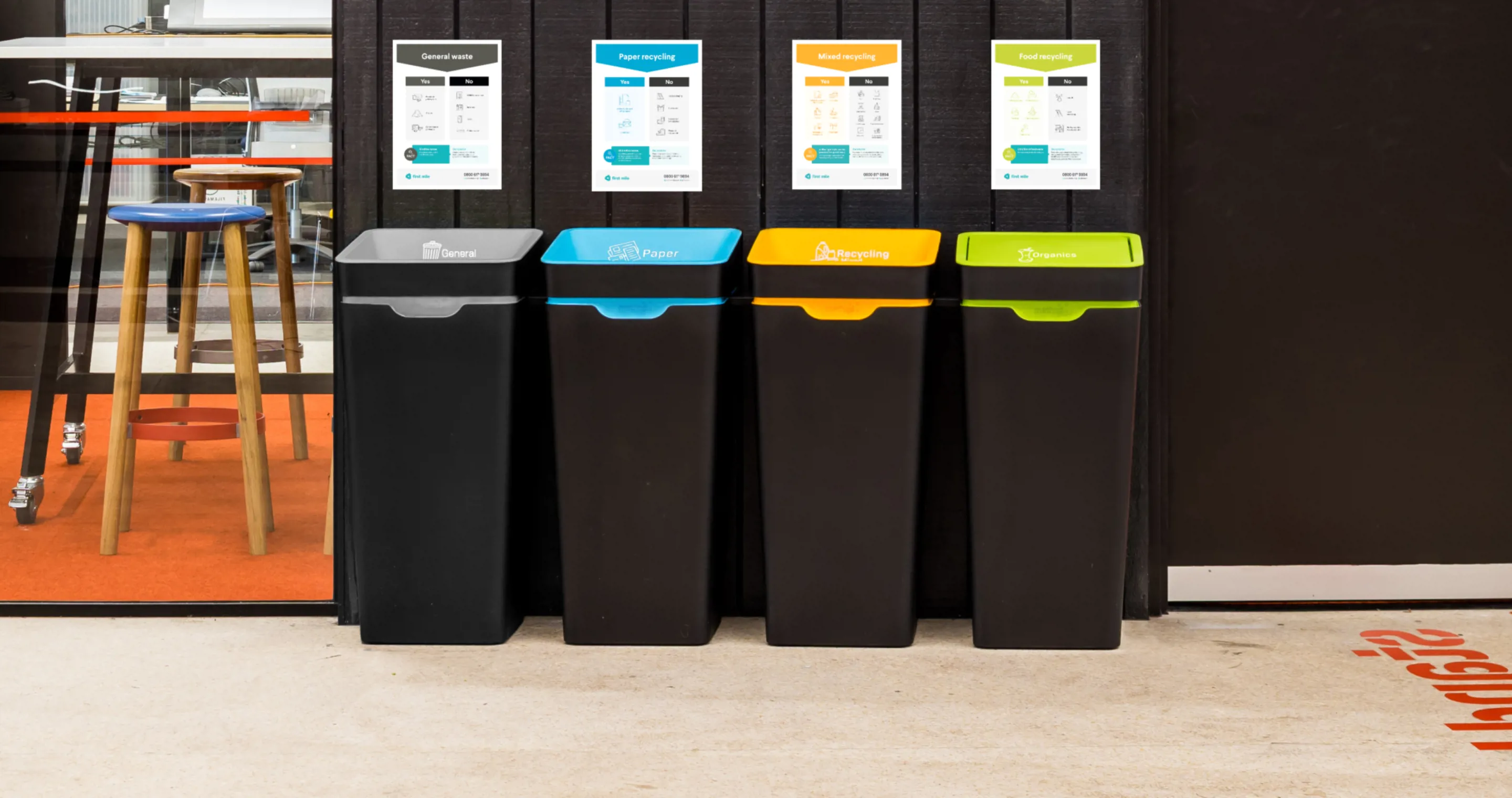 A row of four recycling bins with instructional posters posted above them. The bins are black with different coloured lids.